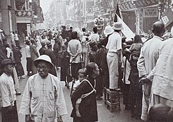 A crowd of people lining a street in Hong Kong watching a parade. Men are visible in a variety of hats.